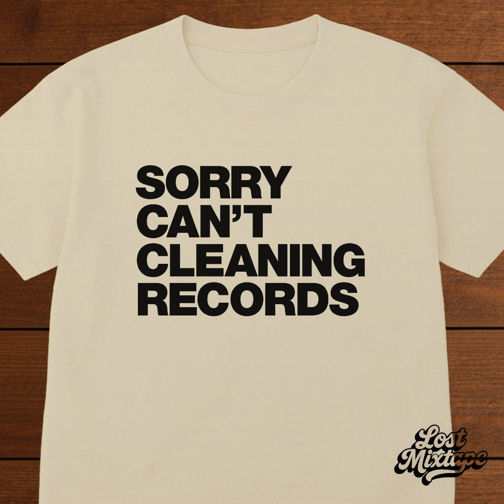 Beige t-shirt with black text 'SORRY CAN'T CLEANING RECORDS' on a wooden surface