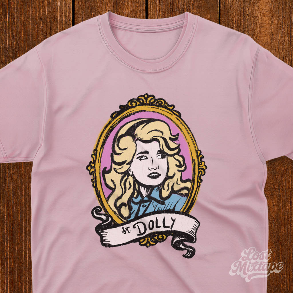 Pink t-shirt with a graphic of a woman's portrait and 'St. Dolly' text on a wooden surface.