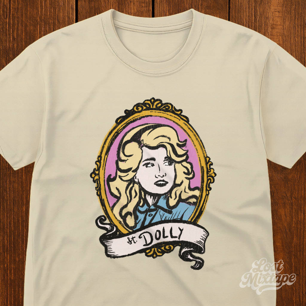 Beige t-shirt with a graphic of a woman's portrait and 'St. Dolly' text on a wooden surface.