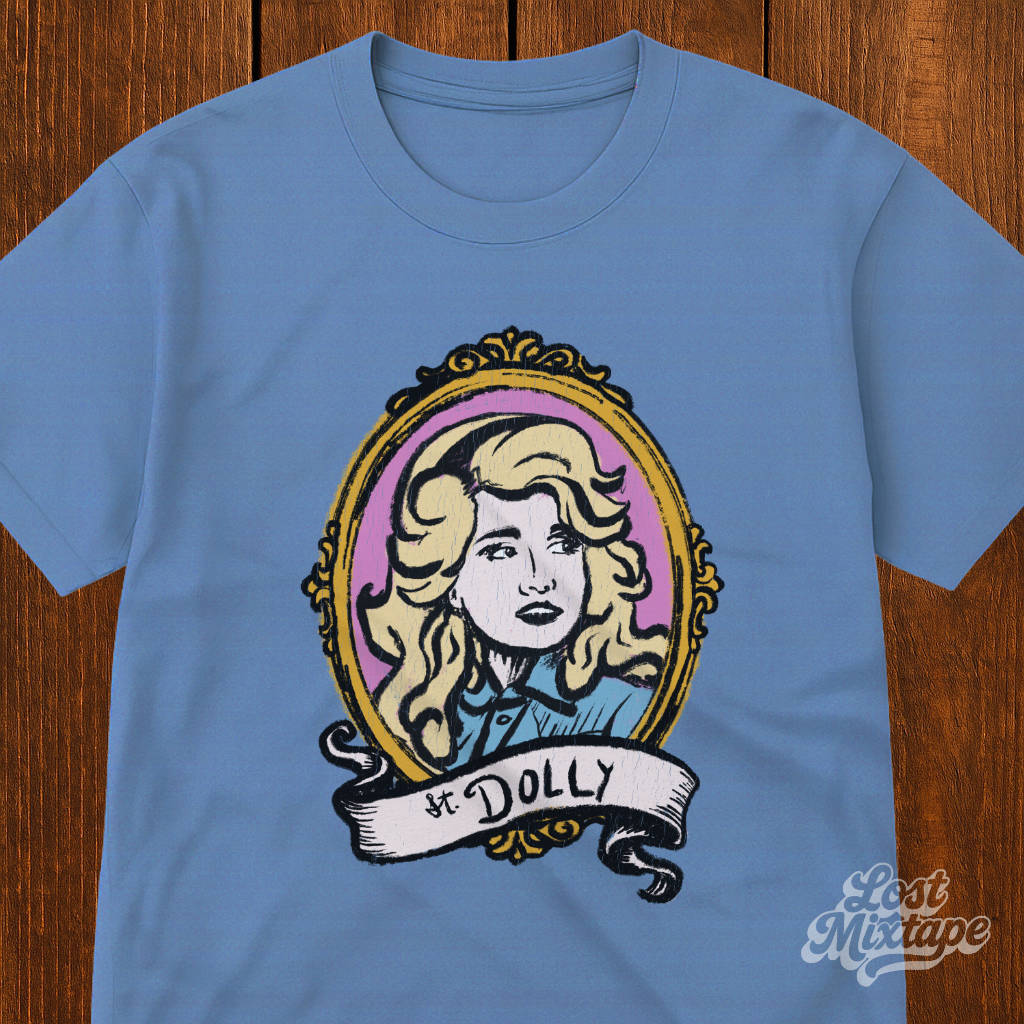 Blue t-shirt with a graphic of a woman's portrait and 'St. Dolly' text on a wooden surface.