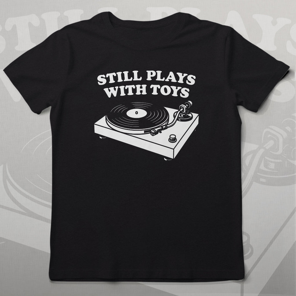 Black t-shirt with a graphic of a turntable and 'Still Plays With Toys' text on a wooden background.